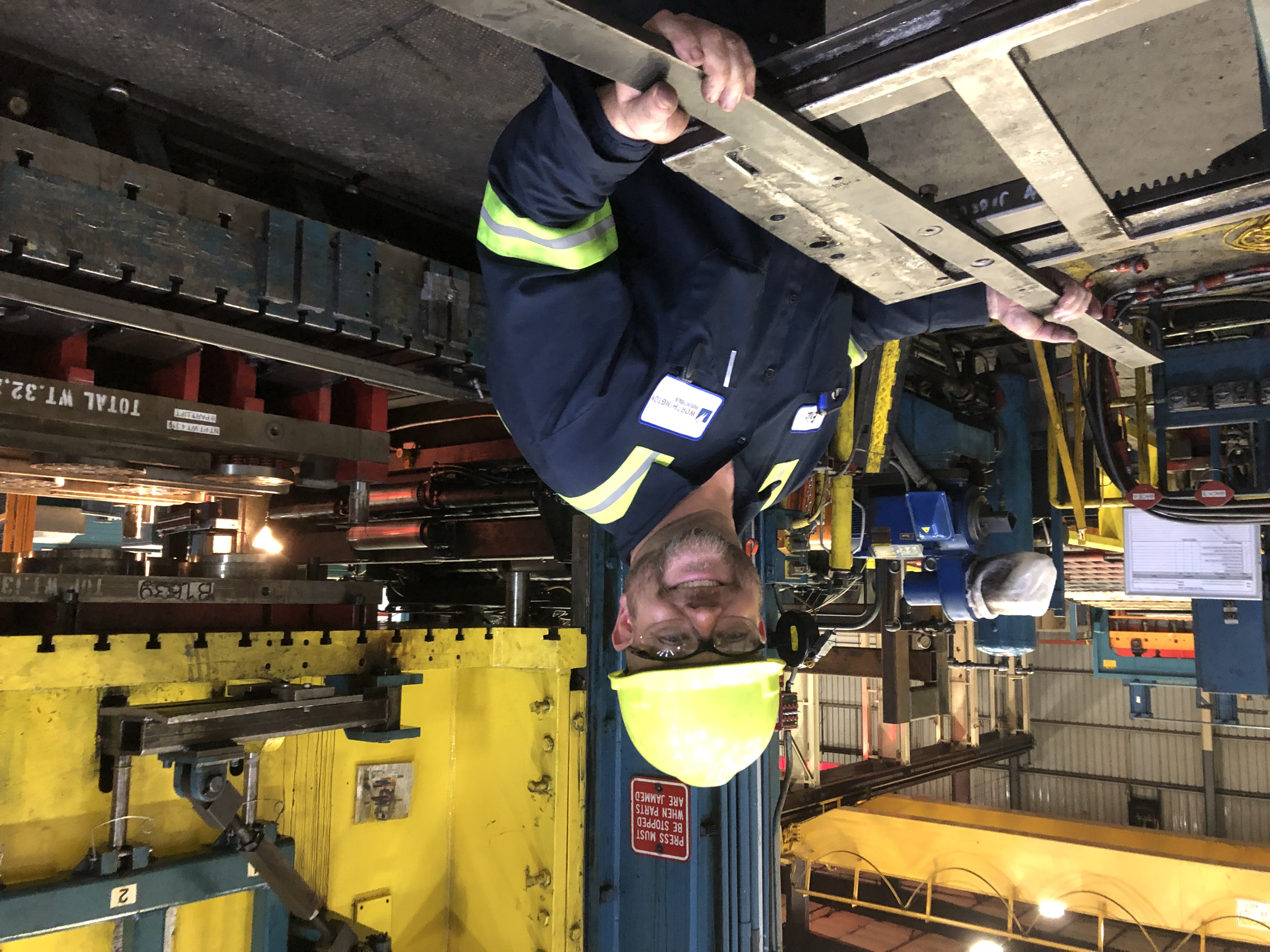 An employee on the manufacturing floor, wearing safety gear, and holding a steel part.